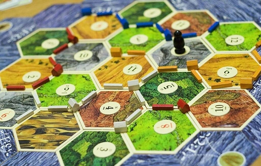 Een opstelling van het bordspel „De Kolonisten van Catan“ met kleurrijke zeshoekige tegels die grondstoffen voorstellen. Verschillende gekleurde wegen en nederzettingen geven de voortgang van de spelers weer