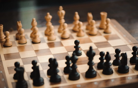 Een schaakbord met houten stukken; zwarte en witte stukken staan tegenover elkaar. Een zwarte pion wordt naar voren geschoven, wat het begin van een strategisch spel suggereert